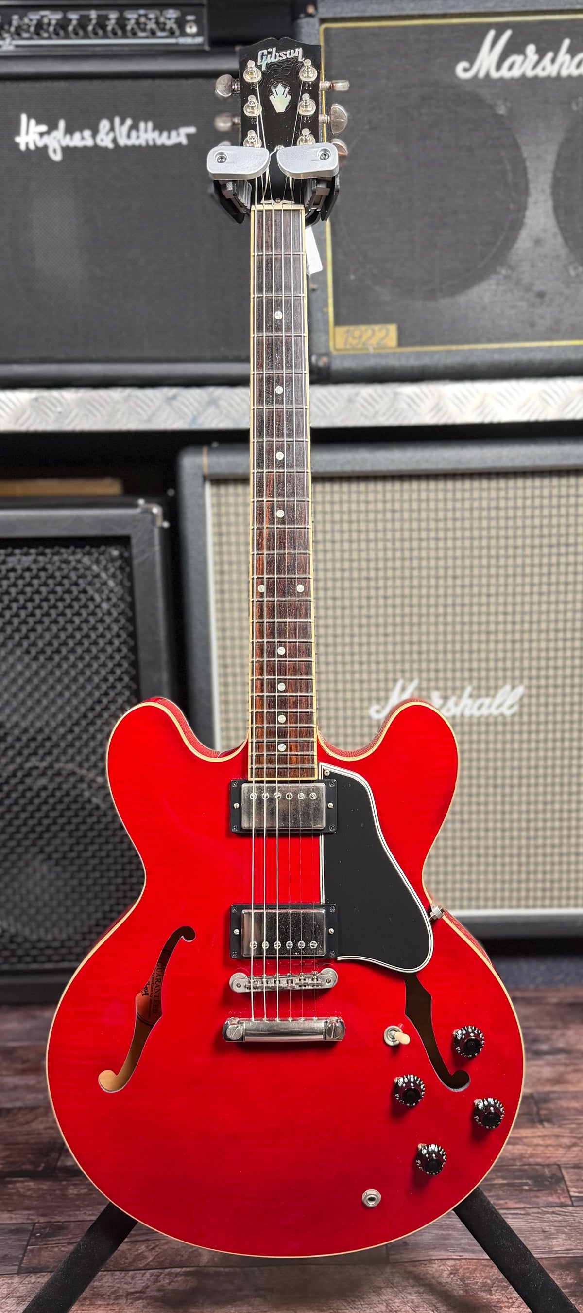 Full-length front view of the guitar upright on a stand, from the Gibson headstock down to the body, set against amp stacks.