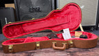 The open, empty case shown from a wider angle with its red plush lining and included accessories laid inside, sitting in front of the shop's amp stacks.