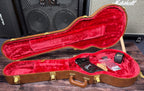 The open, empty case shown from a wider angle with its red plush lining and included accessories laid inside, in front of the shop's amp stacks.