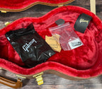Close-up inside the case's accessory pocket, showing a polish cloth bag, plectrums, a Drierite desiccant bag and a Gibson-branded cable or tool tube on the red plush lining.
