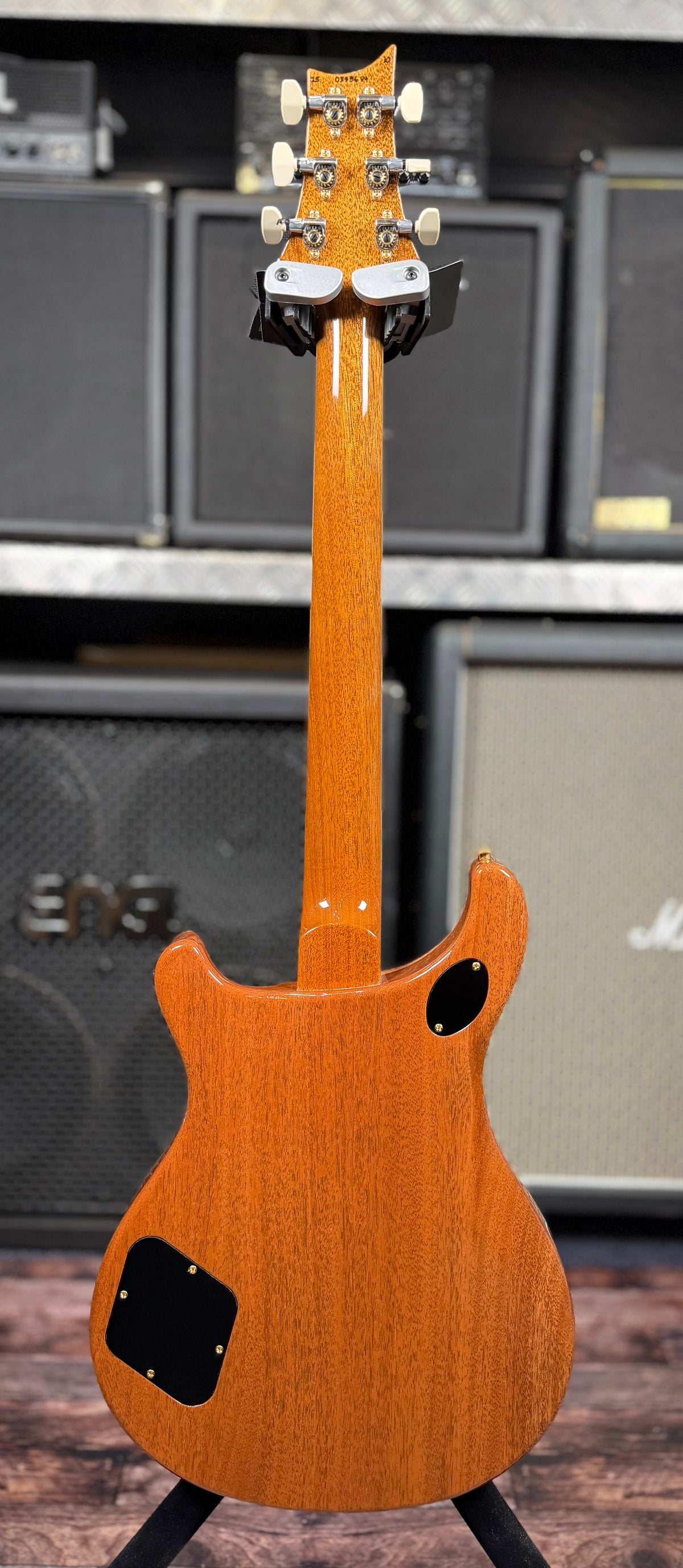 Full-length rear view of the guitar on its stand, showing the mahogany neck, headstock back and body in front of amp stacks.