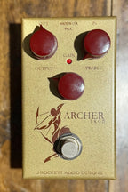 Front view of the J Rockett Audio Designs Archer Ikon pedal in its gold casing with burgundy knobs, showing the archer graphic and Output/Treble controls.