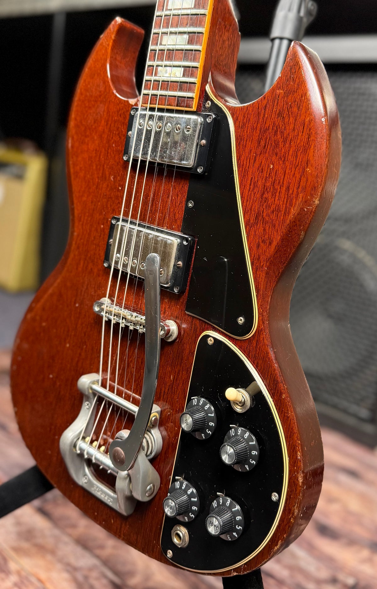 Angled front view of the Gibson SG Deluxe body showing the full control layout of four knobs and a three-way toggle switch, plus the vibrato tailpiece, in front of an amplifier.