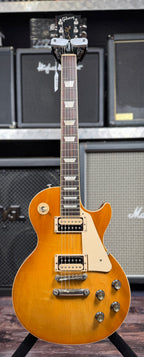 Full front view of the Gibson Les Paul Classic in Honeyburst, showing the Gibson headstock, block fretboard inlays, cream pickguard and single-cutaway body on a stand in front of amp stacks.