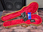 The open case with its red plush lining, showing the Gibson owner's manual, warranty card, polish cloth and spare gold knobs laid inside.