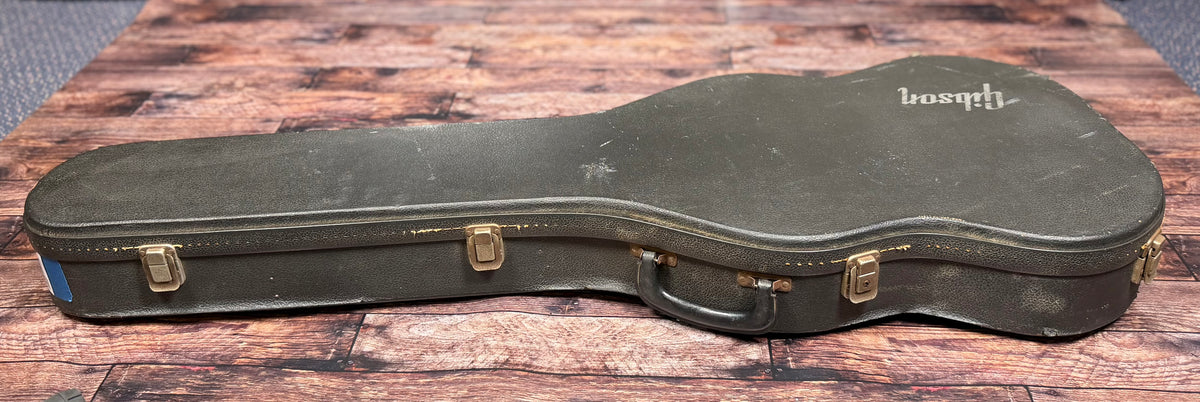 Side view of the closed Gibson hardcase on the floor, showing the latches, carry handle and 'Gibson' logo on the lid.