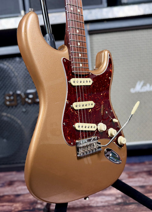 Close-up angled view of the Fender American Professional II Stratocaster's Firemist Gold body and tortoiseshell pickguard, showing the three single-coil pickups, rosewood fretboard edge and tremolo bridge, with amplifiers in the background.